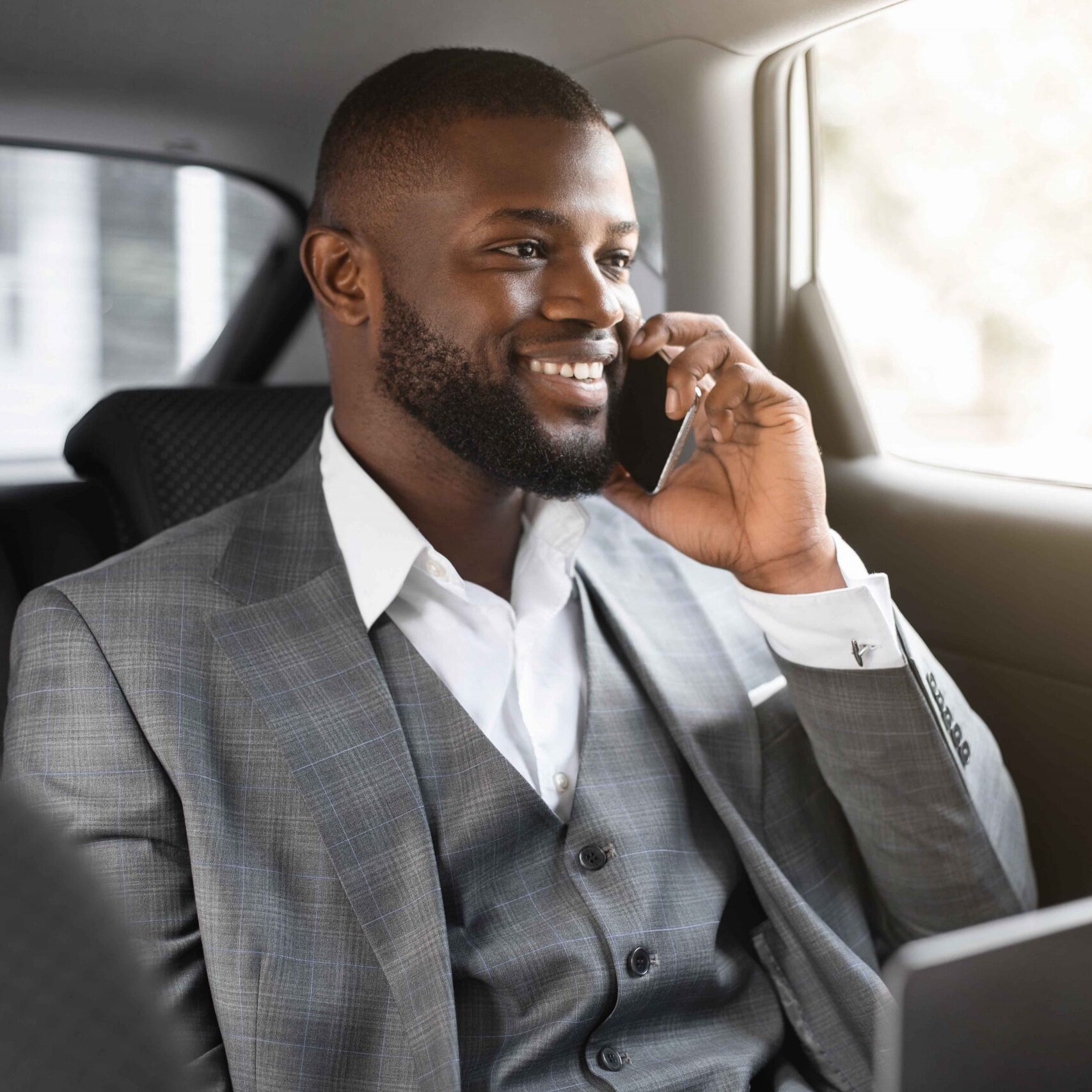 Happy black businessman with laptop talking on mobile phone with clients, looking through window, going to business meeting by car, african manager sitting on back seat, having conversation on phone